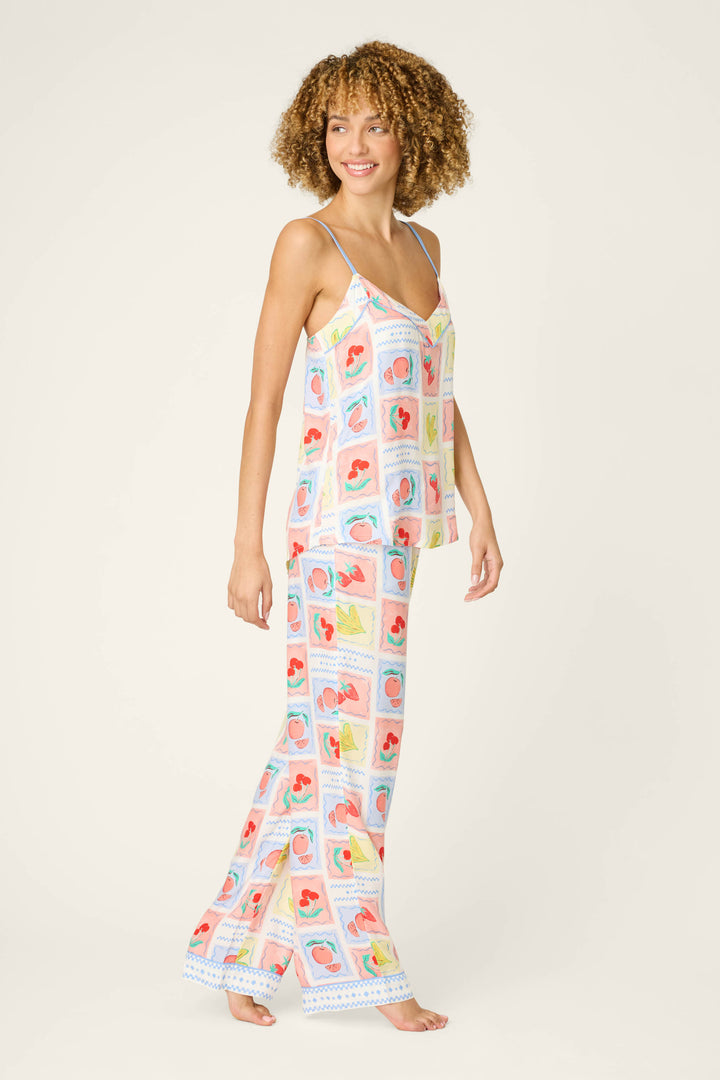 A person with curly hair wears the P.J. Salvage Frutta Fresca Summer Harvest Long Lounge Set, made from silky rayon sateen in a playful multicolor fruit print. They stand barefoot against a plain background.