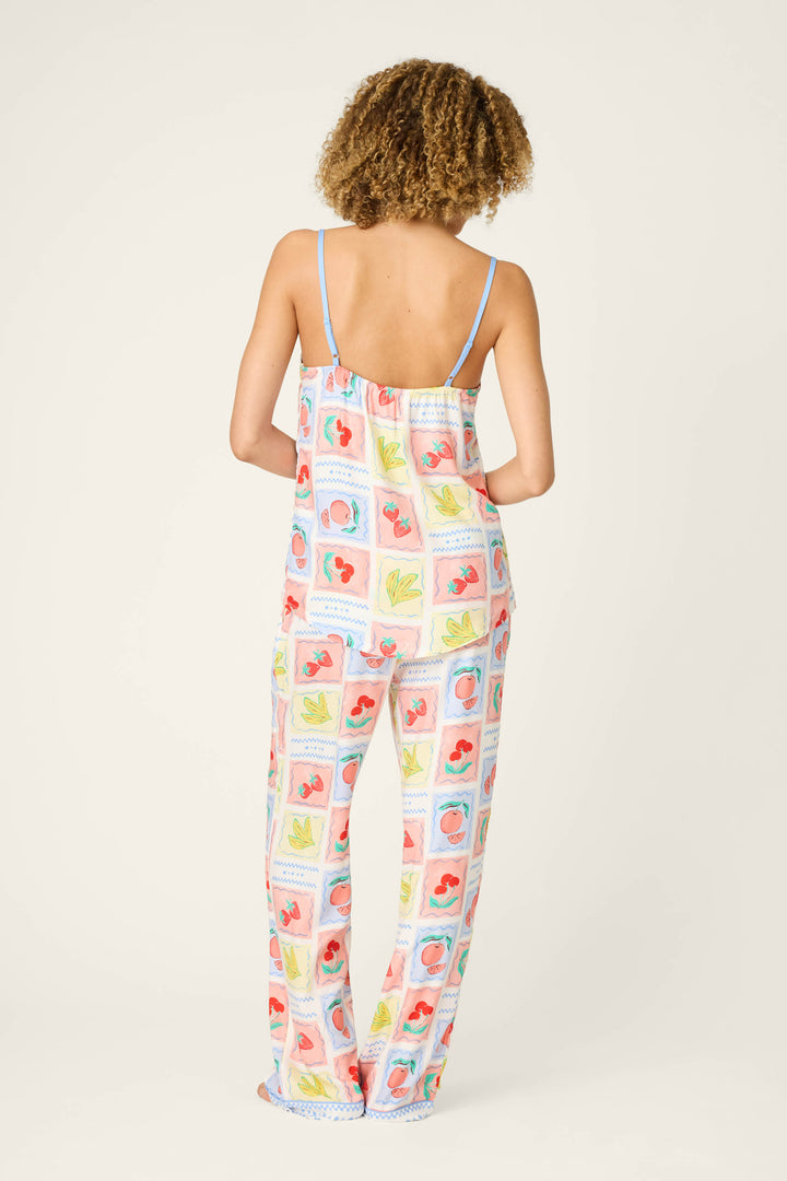 A person with curly hair, seen from behind, wears the Frutta Fresca Summer Harvest Long Lounge Set by P.J. Salvage—a silky rayon sateen pajama set with thin straps and loose pants, featuring colorful square patterns on a white background.