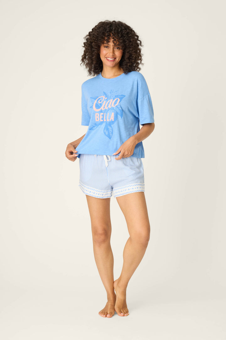 A person with curly hair stands barefoot against a plain background, smiling and holding the side of their blue "Ciao Bella" t-shirt. They wear P.J. Salvage Frutta Fresca Striped Shorts—light blue and white stripes with lace trim—for breathable sleepwear.