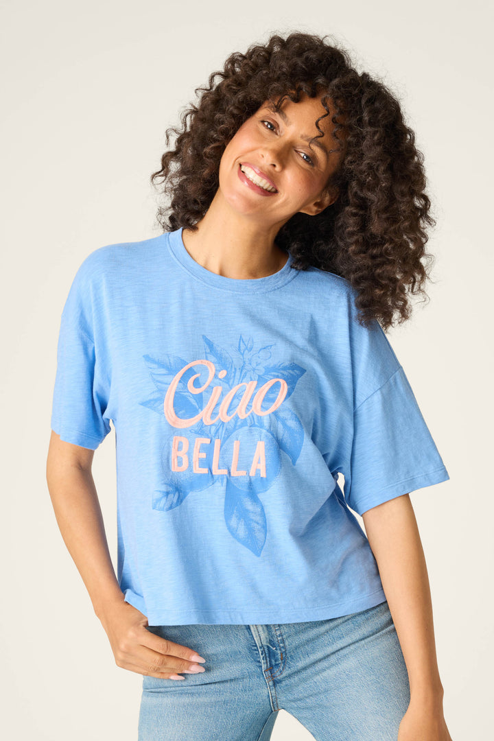 A person with curly hair smiles at the camera, wearing the P.J. Salvage Frutta Fresca Ciao Bella Graphic Tee in light blue, featuring "Ciao Bella" in pink and white letters and a subtle leaf design.