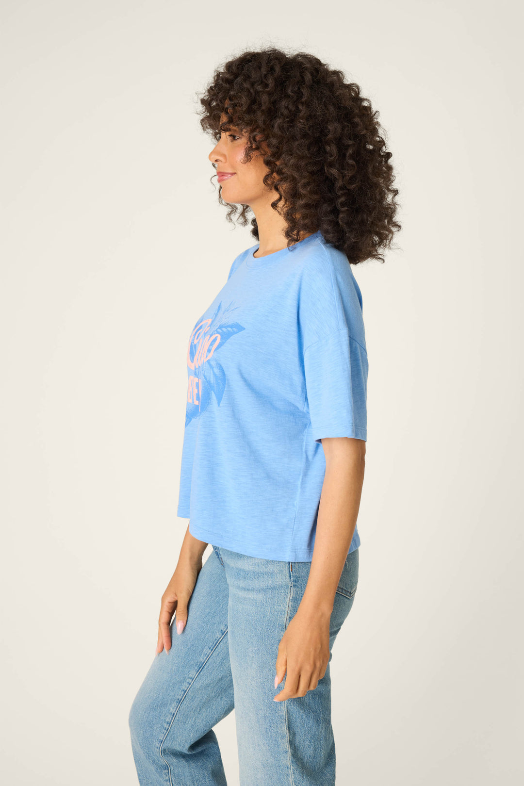 A woman with curly hair stands in profile against a plain background, wearing the P.J. Salvage Frutta Fresca Ciao Bella Graphic Tee in soft light blue cotton-modal jersey and matching light blue jeans, her left hand resting on her thigh.
