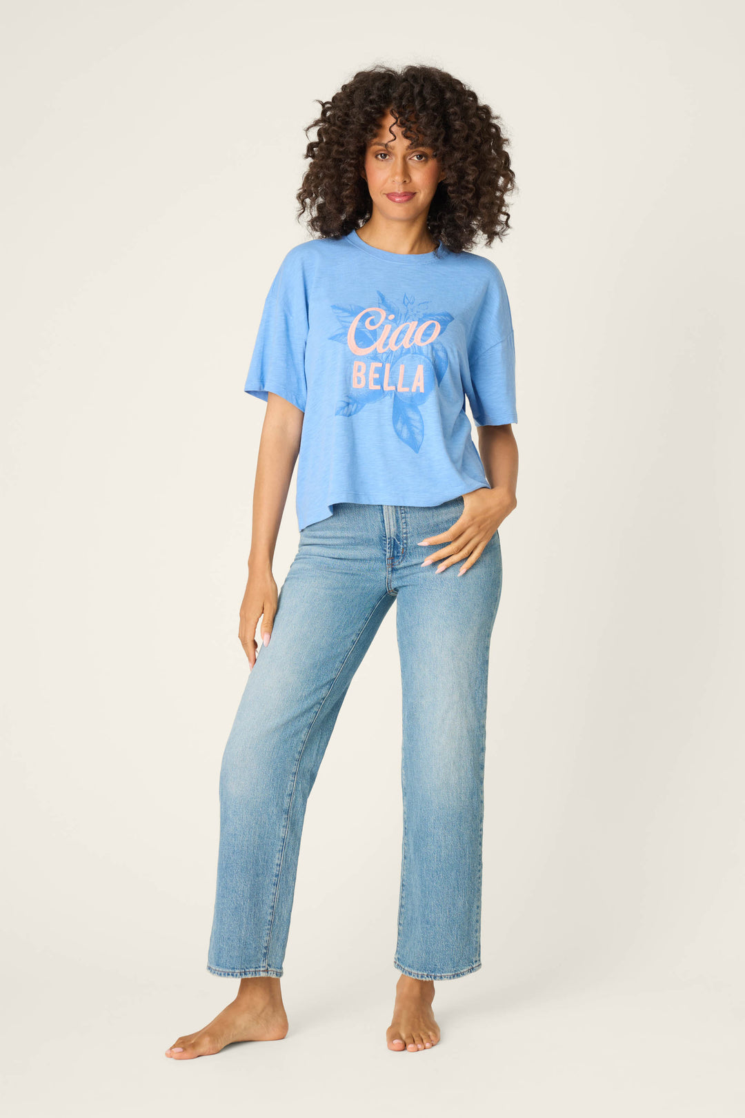 A person with curly hair wears a P.J. Salvage Frutta Fresca Ciao Bella Graphic Tee and matching jeans, barefoot on a light background, one hand on their hip and the other by their side.
