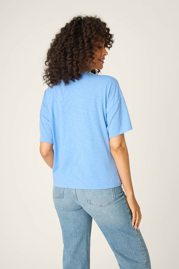 Seen from behind, a person with curly hair wears the P.J. Salvage Frutta Fresca Ciao Bella Graphic Tee in light blue and light blue jeans, standing against a plain light background.