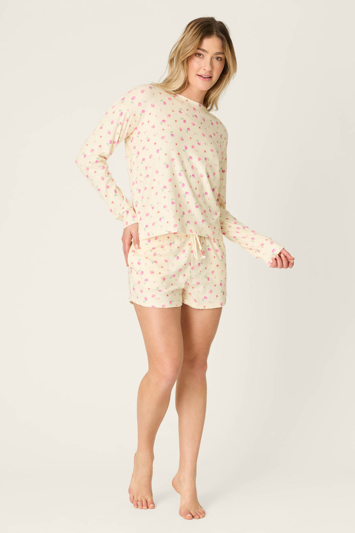 A woman stands barefoot against a plain light background, wearing the P.J. Salvage For the Mamas Butter Blooms Long Sleeve Top in butter yellow with a small pink floral pattern, paired with matching pajama shorts.