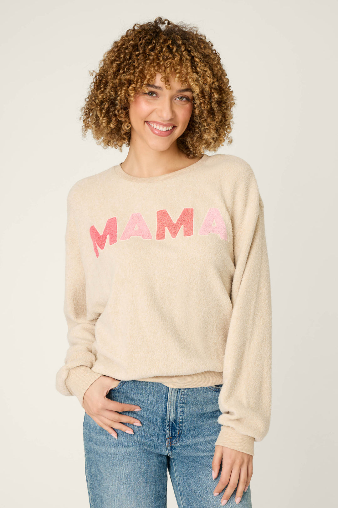 A person with curly hair smiles at the camera, wearing the P.J. Salvage For the Mamas Sweetheart Mama Sweatshirt in light oatmeal, paired with blue jeans, and stands against a plain light background.