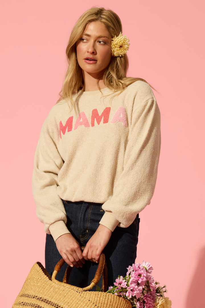 A woman with long blonde hair and a flower behind her ear wears a cream sweatshirt with pink letters, holding flowers on a pink background.