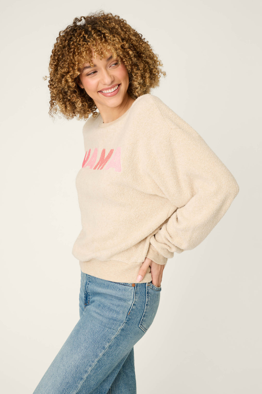 A person with curly hair wears the P.J. Salvage For the Mamas Sweetheart Mama Sweatshirt in light oatmeal, paired with blue jeans. They stand against a plain background, smiling and looking to the side with one hand on their hip.