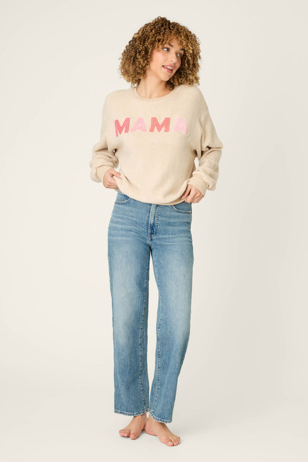 A person with curly hair stands barefoot against a plain background, wearing the P.J. Salvage For the Mamas Sweetheart Mama Sweatshirt in light oatmeal and blue jeans, hands in their pockets and looking to the side.