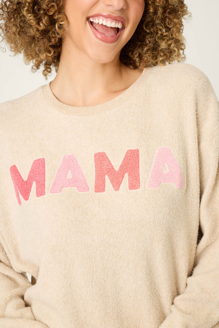 A woman with curly hair smiles, wearing the P.J. Salvage For the Mamas Sweetheart Mama Sweatshirt in cozy cloud brushed terry. The light oatmeal long sleeve features large pink and red 'MAMA' letters. Only her mouth and upper torso are visible.