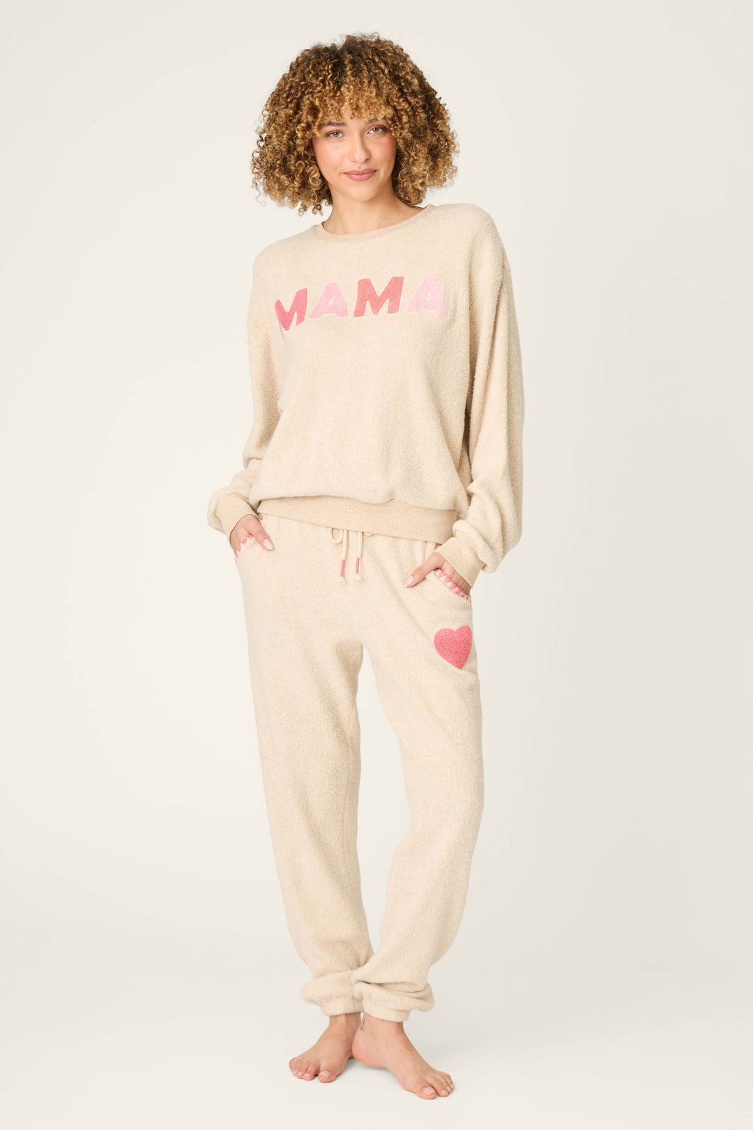 A person with curly hair wears the P.J. Salvage For the Mamas Sweetheart Banded Pants, featuring a pink heart, with a beige top reading MAMA in pink letters. They stand barefoot against a plain light background.