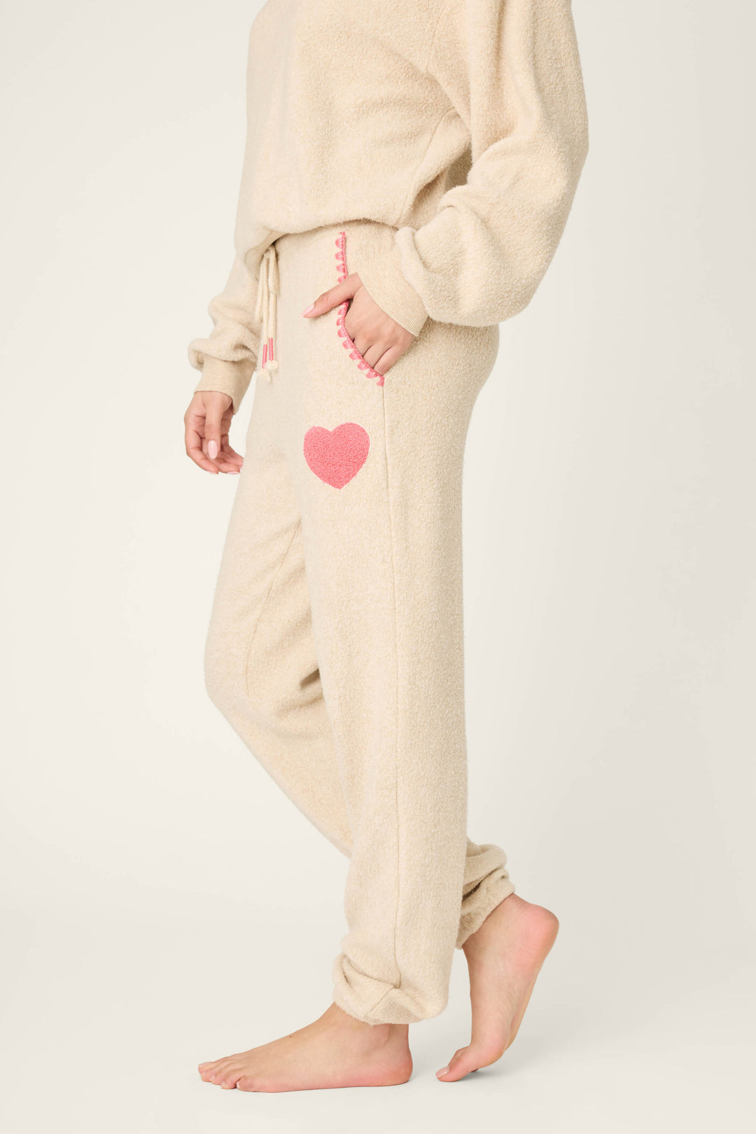 A person stands barefoot in P.J. Salvage For the Mamas Sweetheart Banded Pants—beige, fuzzy lounge pants with a pink heart on the pocket and pink stitching—showcasing their cozy plushness and a partially visible matching top.