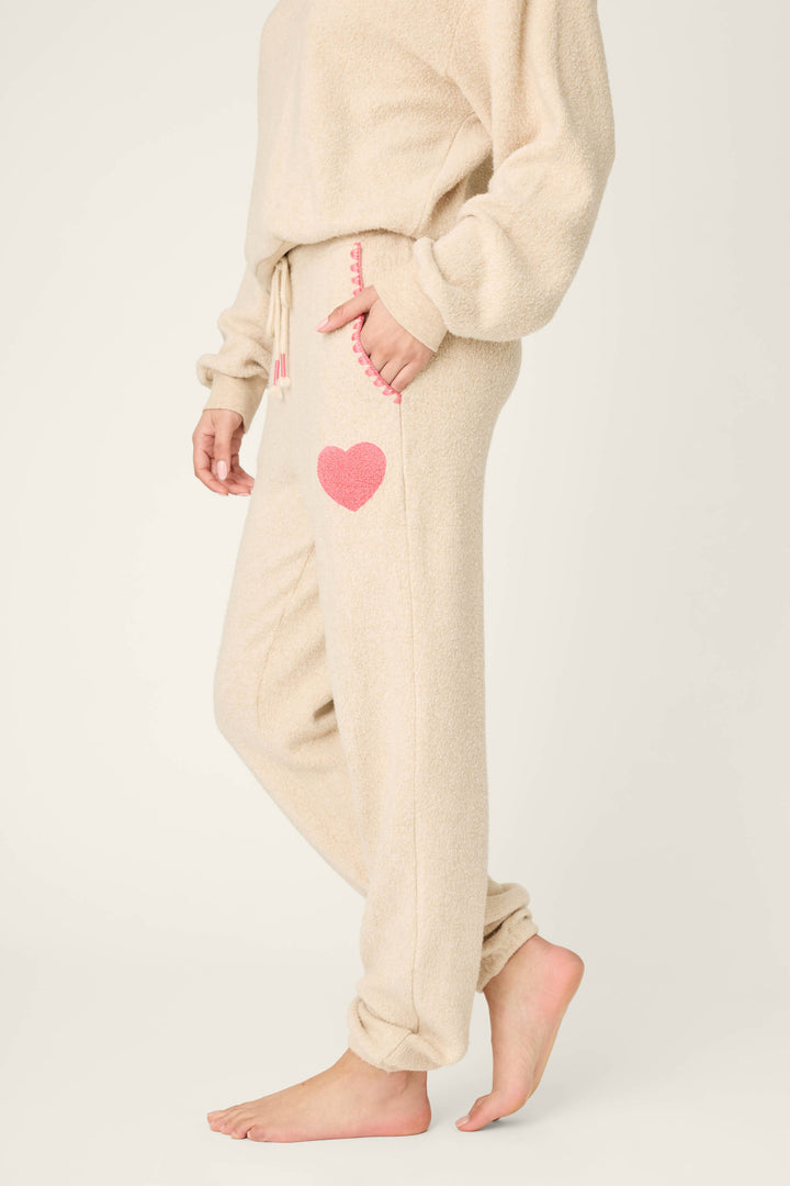 A person stands barefoot in P.J. Salvage For the Mamas Sweetheart Banded Pants—beige, fuzzy lounge pants with a pink heart on the pocket and pink stitching—showcasing their cozy plushness and a partially visible matching top.