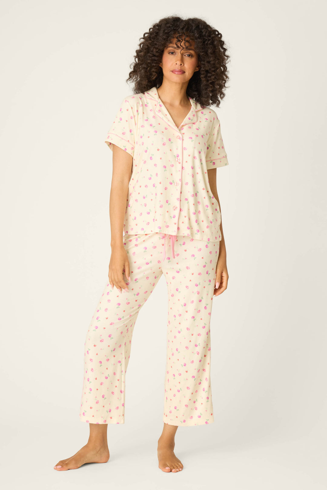 A woman with curly hair and a neutral expression wears the P.J. Salvage For the Mamas Butter Blooms Cropped 3-Piece PJ Set—barefoot in cozy butter yellow pointelle pajamas with a small pink pattern, featuring short sleeves and long pants.