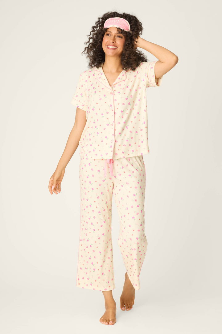 A woman smiles barefoot against a plain background, wearing P.J. Salvage's For the Mamas Butter Blooms Cropped 3-Piece PJ Set—cream pajamas with pink florals and a matching pink sleep mask, one hand resting on her mask.