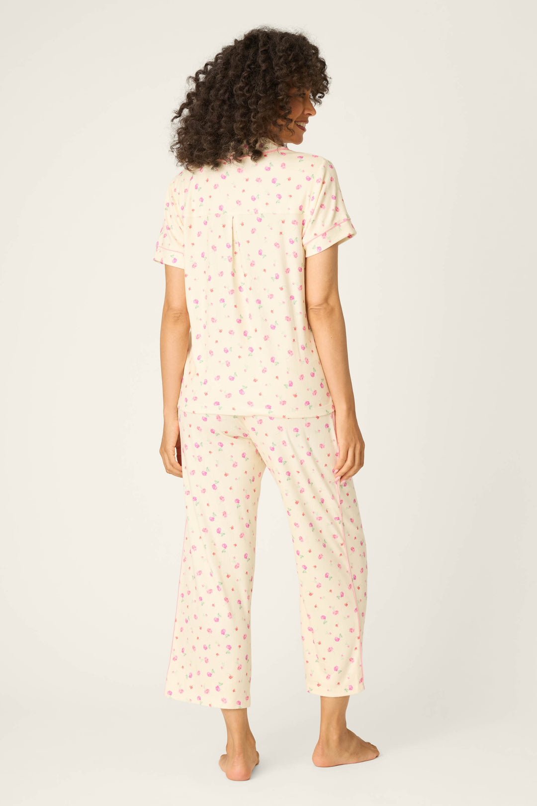 A woman faces away, barefoot, in the P.J. Salvage For the Mamas Butter Blooms Cropped 3-Piece PJ Set—short sleeves and loose full-length pants in a light cream with pink florals—set against a plain off-white background for a relaxed vibe.