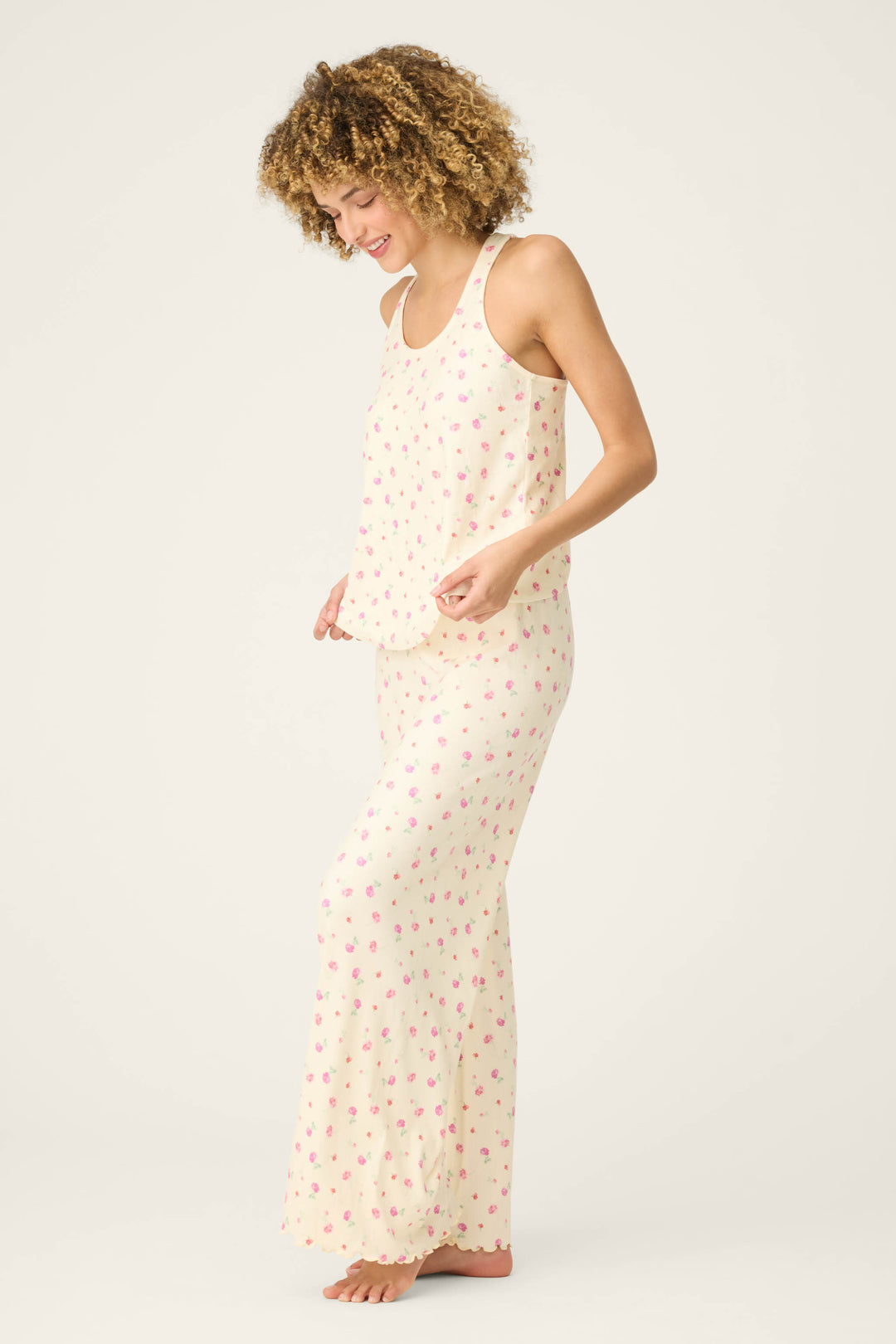 A person stands barefoot on a light background, smiling and gently holding the hem of their For the Mamas Butter Blooms Long PJ Set—a sleeveless, cream-colored floral pajama set by P.J. Salvage.