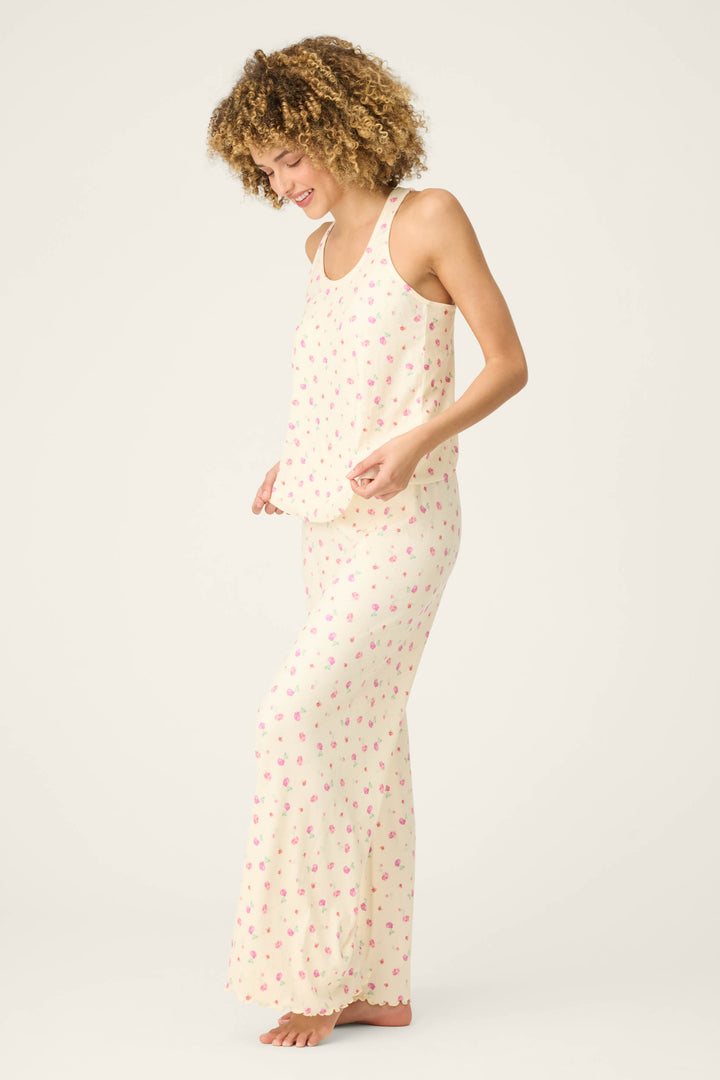 A person stands barefoot on a light background, smiling and gently holding the hem of their For the Mamas Butter Blooms Long PJ Set—a sleeveless, cream-colored floral pajama set by P.J. Salvage.