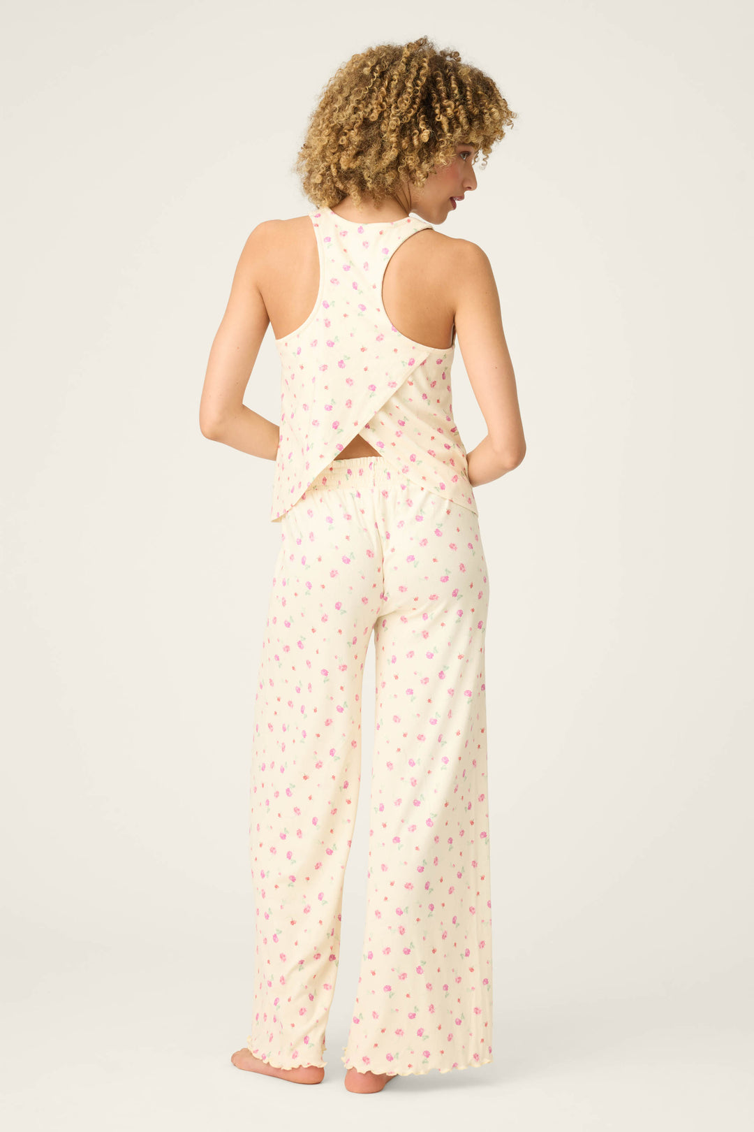 A person stands barefoot facing away, wearing the P.J. Salvage For the Mamas Butter Blooms Long PJ Set, a light-colored cross-back floral pajama top and matching wide-leg pants. The background is plain and neutral.
