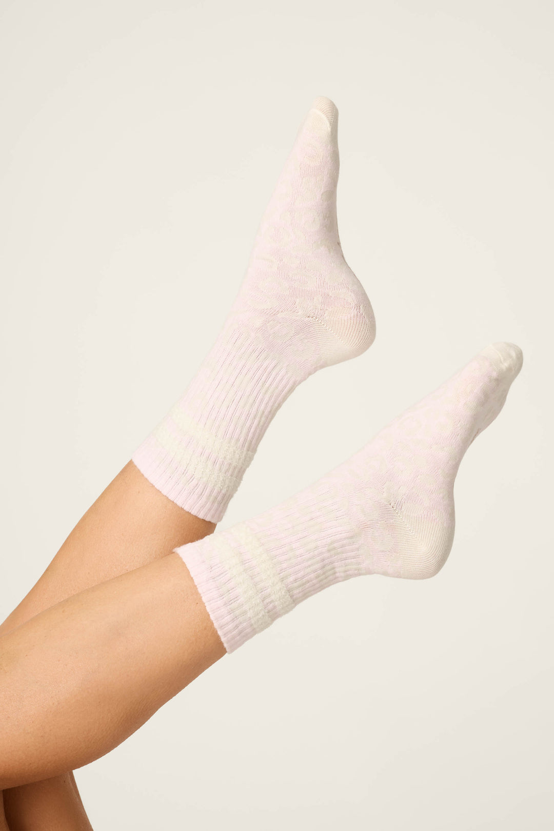 A pair of feet in P.J. Salvage's Luxe Leopard Dreams Ribbed Crew Socks, shown in ballet-pink with a subtle pattern, set against an off-white background and legs raised, crossed at the ankles.