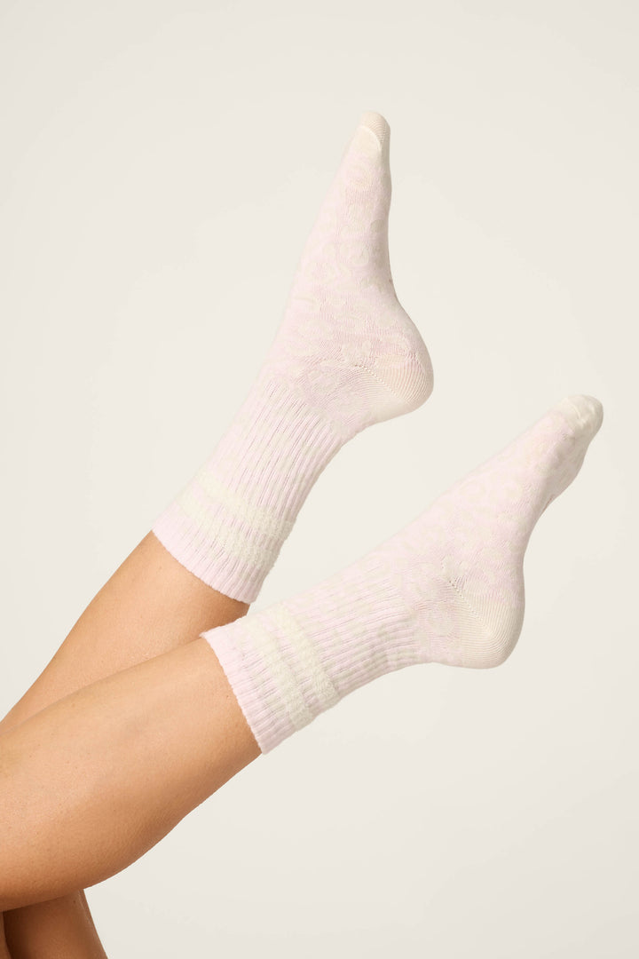 A pair of feet in P.J. Salvage's Luxe Leopard Dreams Ribbed Crew Socks, shown in ballet-pink with a subtle pattern, set against an off-white background and legs raised, crossed at the ankles.