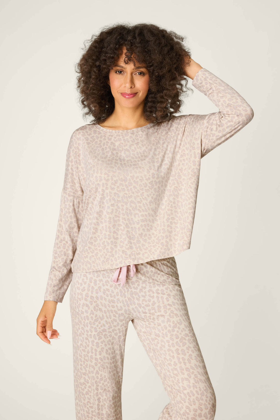 A person with curly hair wears the P.J. Salvage Luxe Leopard Dreams Long Sleeve Top, featuring a soft jersey fabric and light leopard print, paired with matching pants, standing against a simple light-colored background.