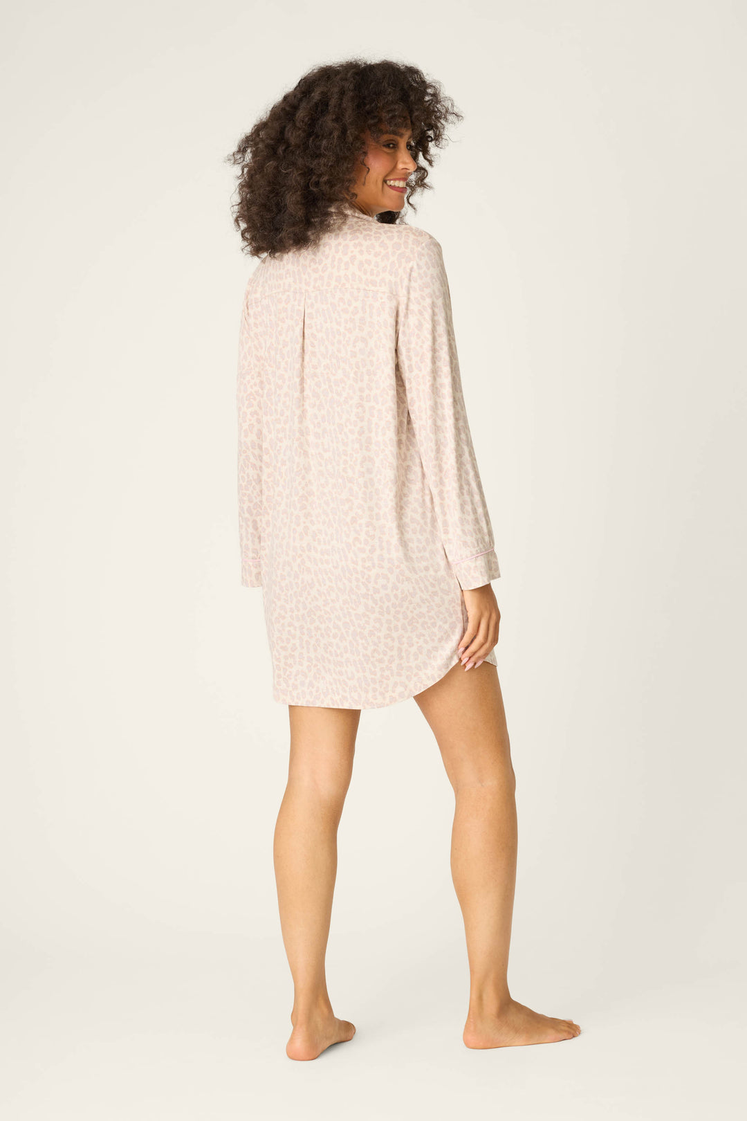 A person with curly hair stands barefoot, turning to smile at the camera. They wear the P.J. Salvage Luxe Leopard Dreams Nightshirt—a soft, button-front jersey with a subtle leopard print and knee-length silhouette against a plain background.