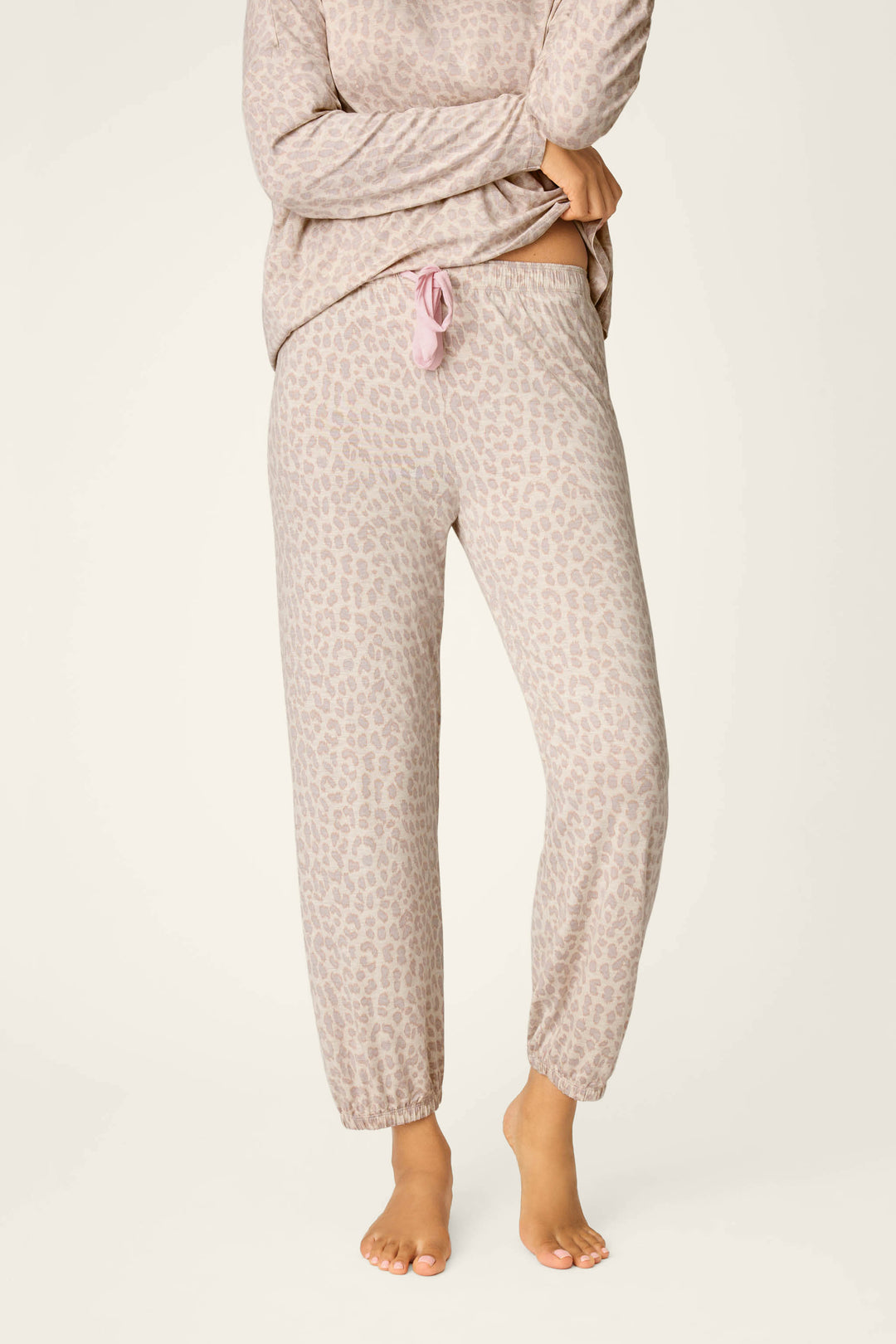 A person stands barefoot in P.J. Salvage Luxe Leopard Dreams Banded Pants, a soft jersey lounge set with long sleeves, featuring light beige and taupe animal print, elastic waistband, and pink drawstring.