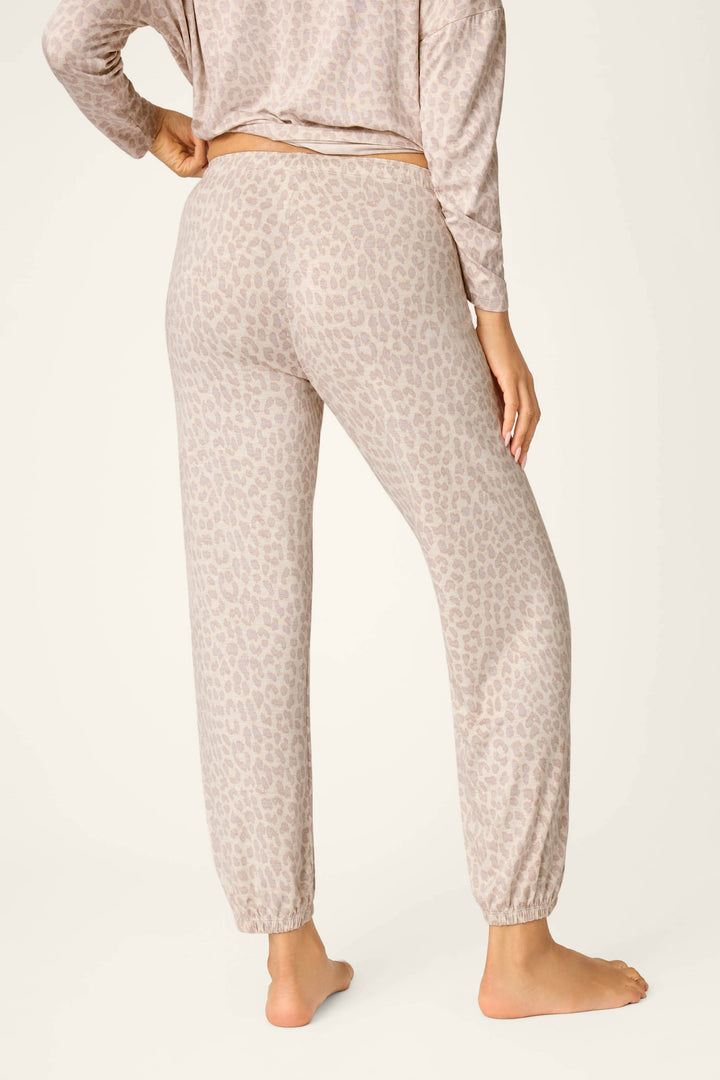 A person stands barefoot facing away, wearing P.J. Salvage Luxe Leopard Dreams Banded Pants—loose-fit leopard lounge pants in soft jersey with a subtle print, elastic waistband, and cuffs.