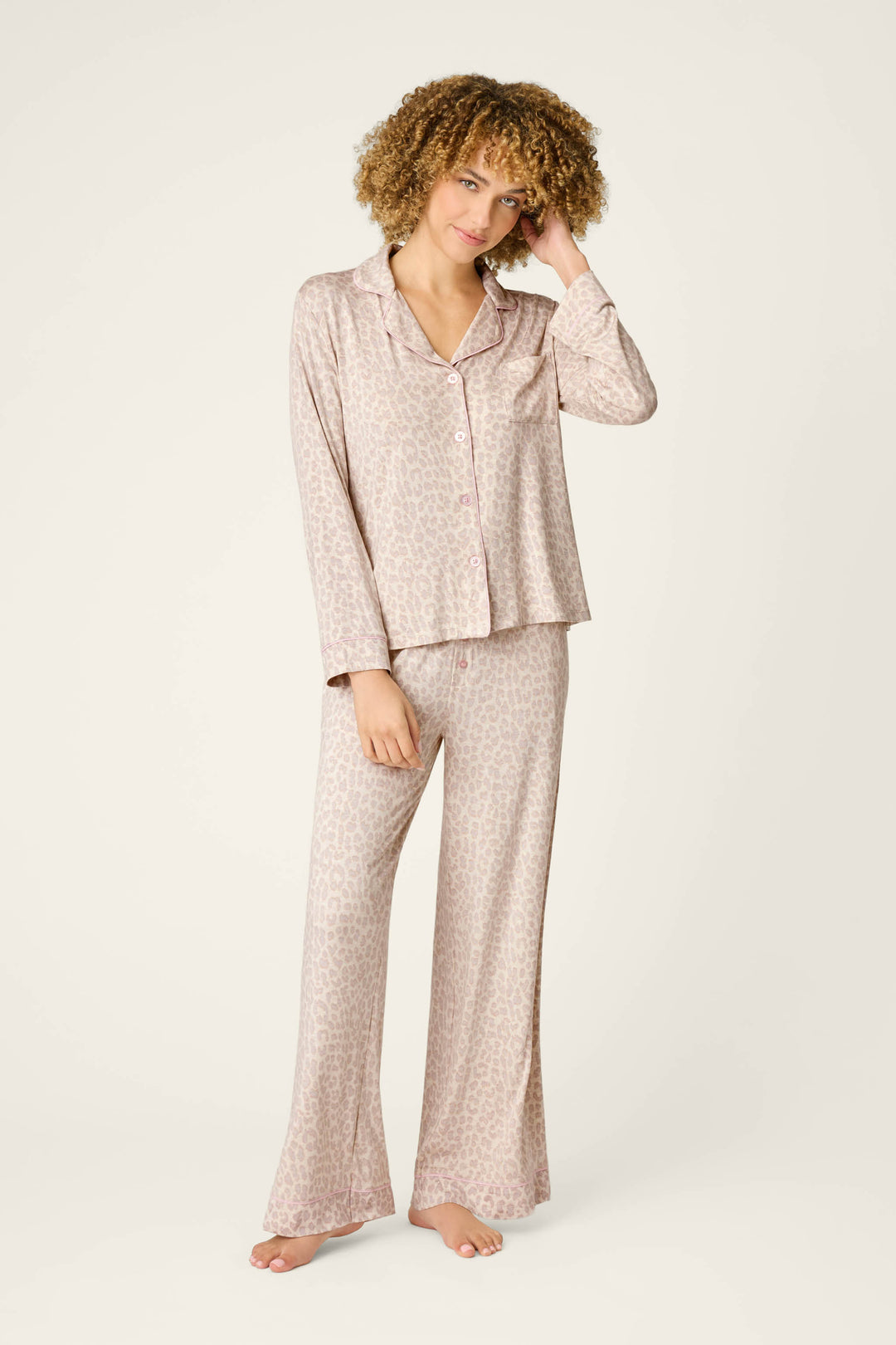 A person with curly hair models the P.J. Salvage Luxe Leopard Dreams PJ Set—a light pink leopard print jersey ensemble with a long-sleeve button-up top and wide-leg pants—standing barefoot against a plain light background.