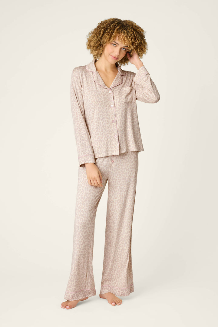 A person with curly hair models the P.J. Salvage Luxe Leopard Dreams PJ Set—a light pink leopard print jersey ensemble with a long-sleeve button-up top and wide-leg pants—standing barefoot against a plain light background.