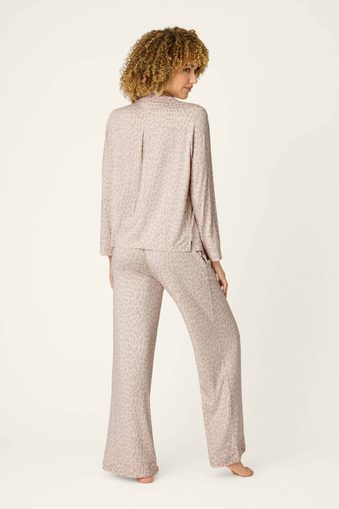A person with curly hair stands barefoot, facing away, wearing the Luxe Leopard Dreams PJ Set by P.J. Salvage—featuring a loose soft jersey top and wide-leg pants in a light leopard pattern—against a plain light background.