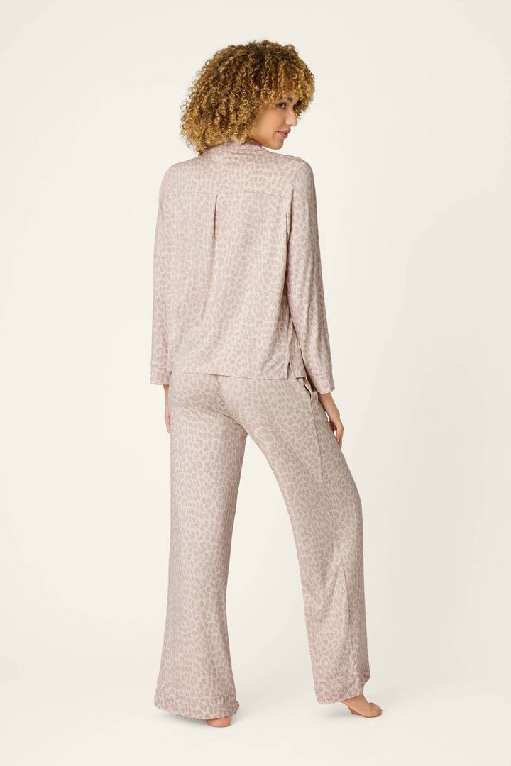 A person with curly hair stands barefoot, facing away, wearing the Luxe Leopard Dreams PJ Set by P.J. Salvage—featuring a loose soft jersey top and wide-leg pants in a light leopard pattern—against a plain light background.