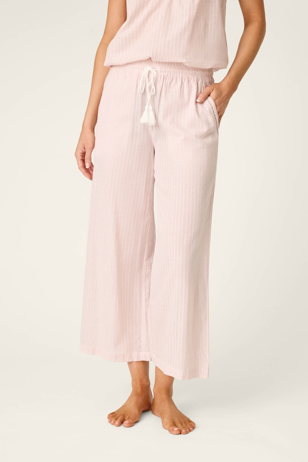 A person stands barefoot in P.J. Salvage On Cloud Nine Silver Rose Open Leg Pajama Pants with subtle vertical stripes, an elastic waistband, drawstring tie, and pockets, paired with a matching top for a relaxed look.