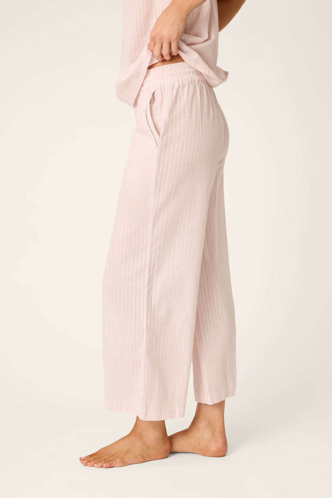 A person stands barefoot on a light surface, wearing P.J. Salvage’s On Cloud Nine Silver Rose Open Leg Pajama Pants—light pink, wide-leg, cropped and striped cotton lounge pants with a matching top, hand in the pocket.