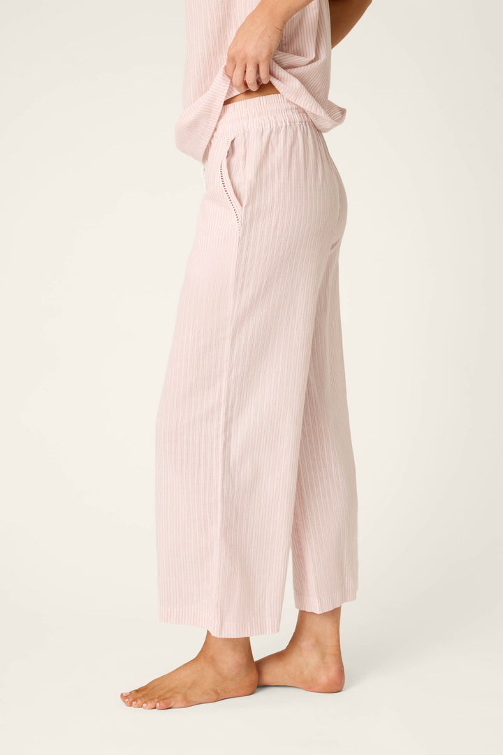 A person stands barefoot on a light surface, wearing P.J. Salvage’s On Cloud Nine Silver Rose Open Leg Pajama Pants—light pink, wide-leg, cropped and striped cotton lounge pants with a matching top, hand in the pocket.