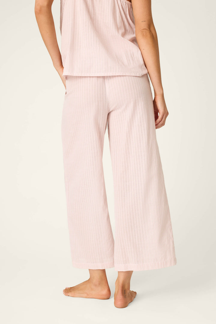 A person stands barefoot, shown from the back, wearing P.J. Salvage On Cloud Nine Silver Rose Open Leg Pajama Pants in light pink with a matching sleeveless top featuring subtle vertical stripes.