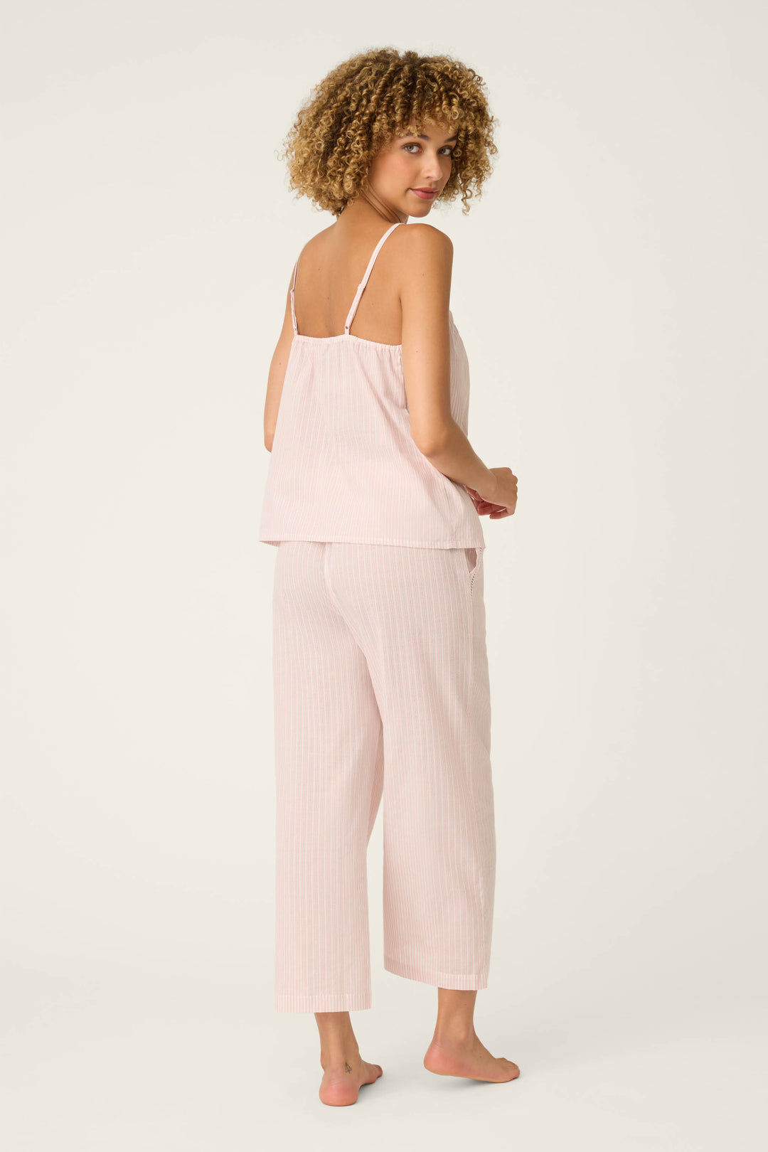 A person with curly hair stands barefoot, facing backward and looking over their shoulder, wearing the P.J. Salvage On Cloud Nine Silver Rose Long PJ Set— a light pink, loose-fitting sleeveless top and matching wide-leg pants—against a plain background.