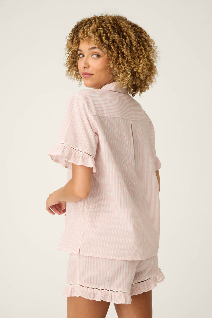 A person with curly hair is standing on a plain background, wearing P.J. Salvage’s On Cloud Nine Silver Rose Short Sleeve Pajama Top and matching shorts, both with ruffle trim and light pink stripes, looking over their shoulder.