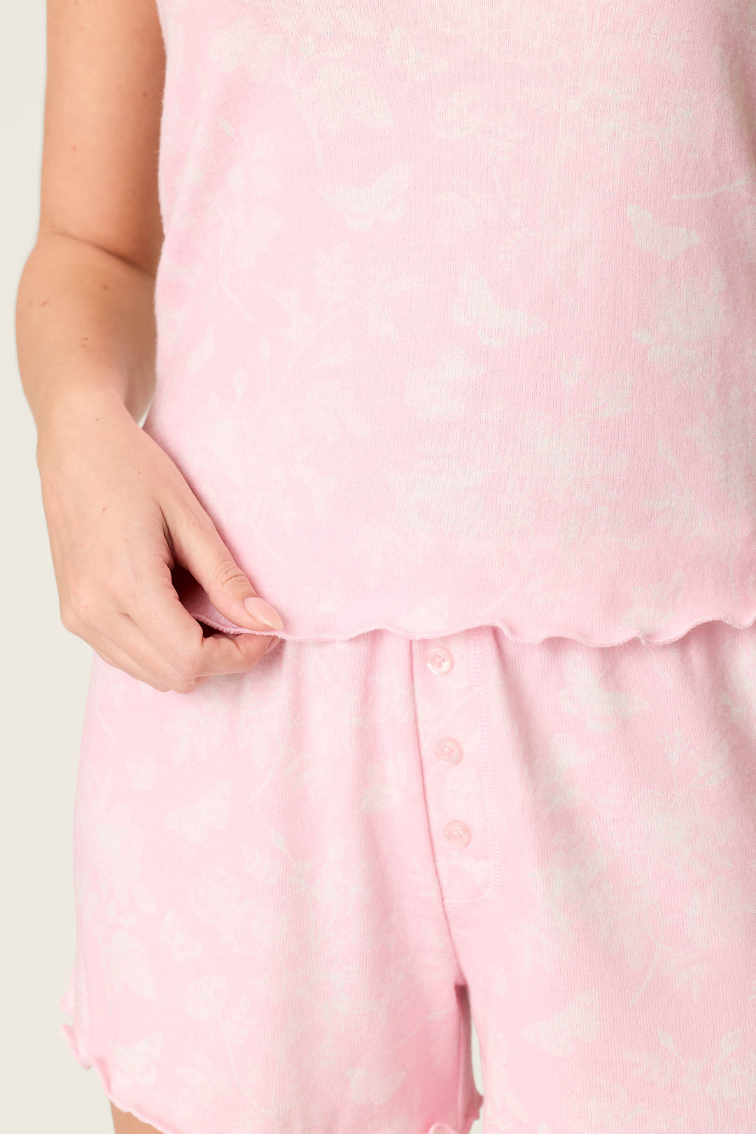 A person wears the P.J. Salvage Peachy Mix & Match Butterfly Blossoms Tank Top in baby pink with a subtle butterfly print, paired with matching buttoned shorts. The peachy jersey fabric adds comfort against a light, plain background.