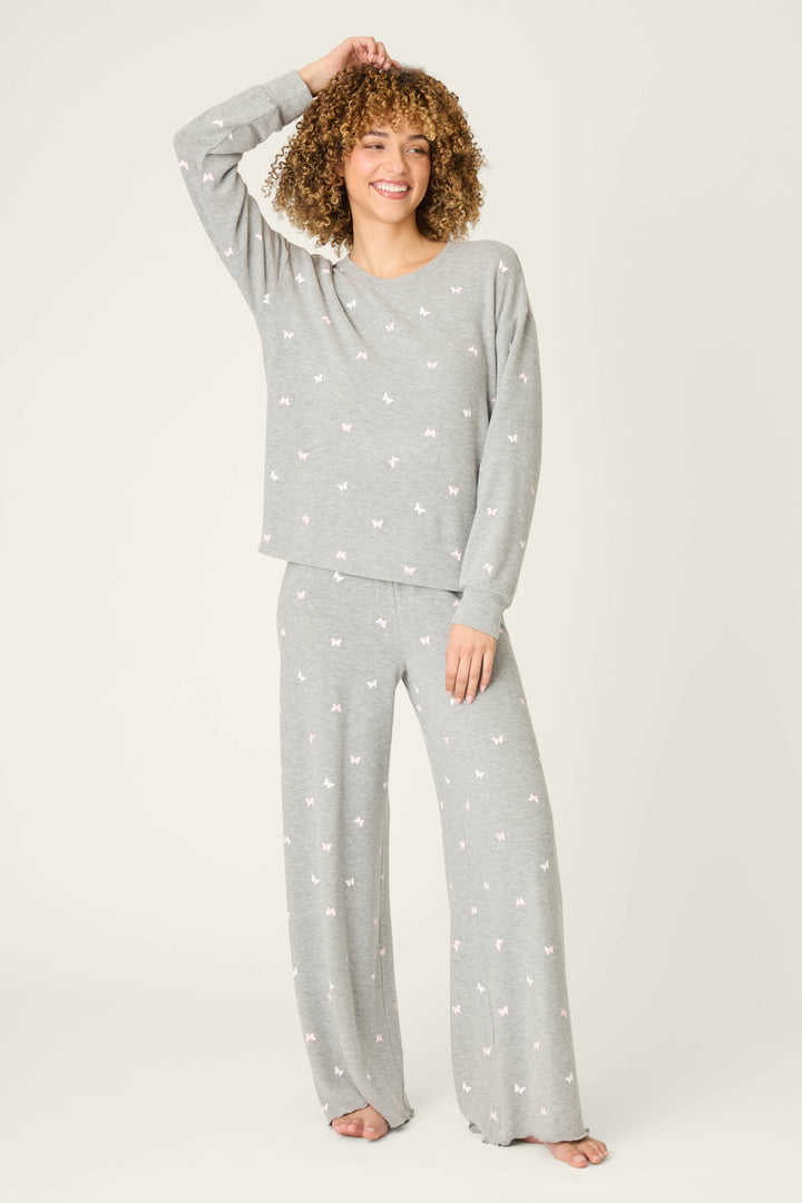 A person stands barefoot against a light background, smiling with one hand raised to their head, wearing the P.J. Salvage Peachy Mix & Match Butterfly Kisses Long Sleeve Top in heather grey with a subtle butterfly print.