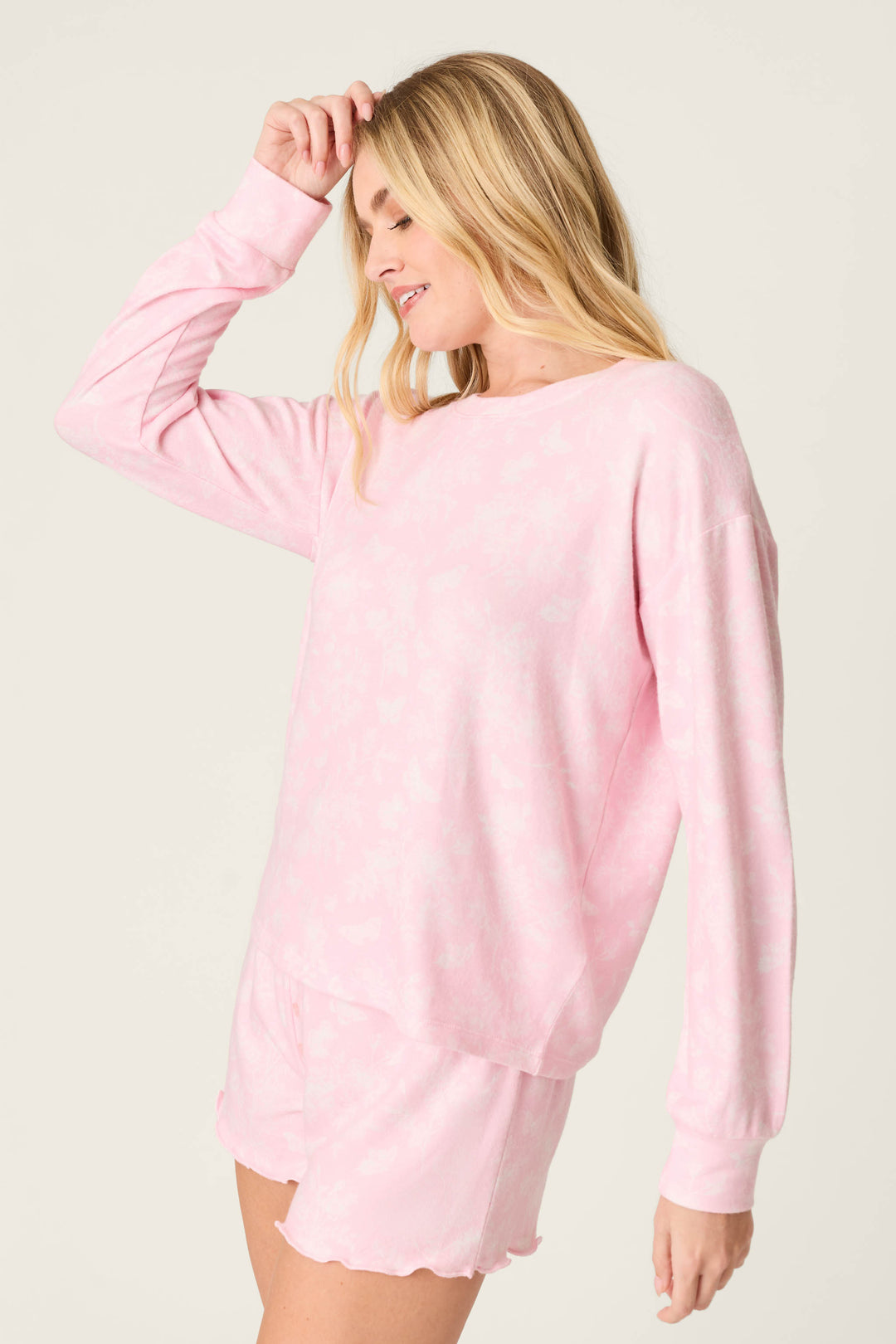 A woman with long blonde hair wears the P.J. Salvage Peachy Mix & Match Butterfly Blossoms Long Sleeve Top in baby pink, paired with matching shorts. She stands sideways, smiling with one hand near her head against a plain, light background.