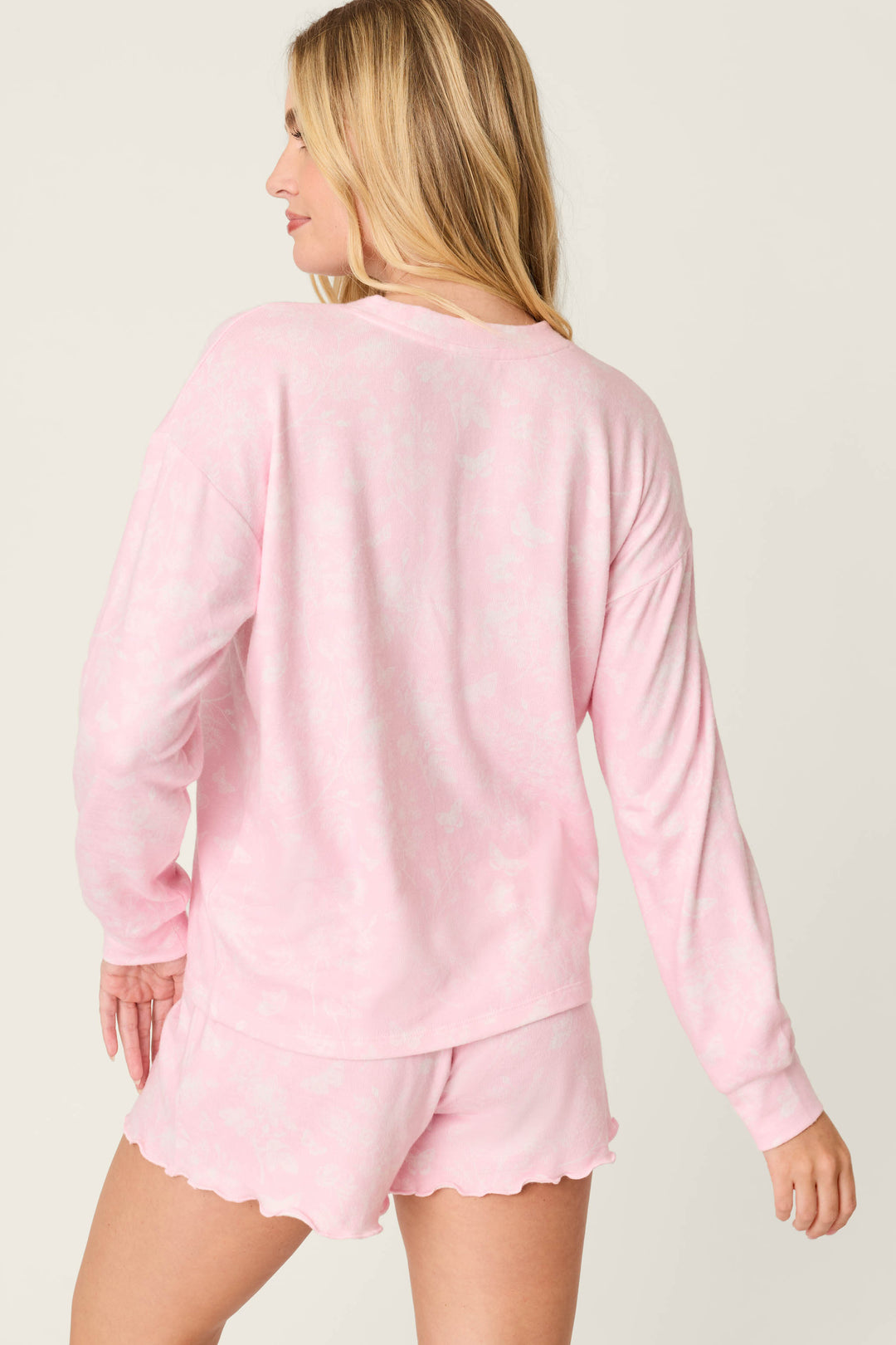 A person with long blonde hair is seen from the back wearing the P.J. Salvage Peachy Mix & Match Butterfly Blossoms Long Sleeve Top in baby pink, paired with matching shorts, standing against a neutral background.