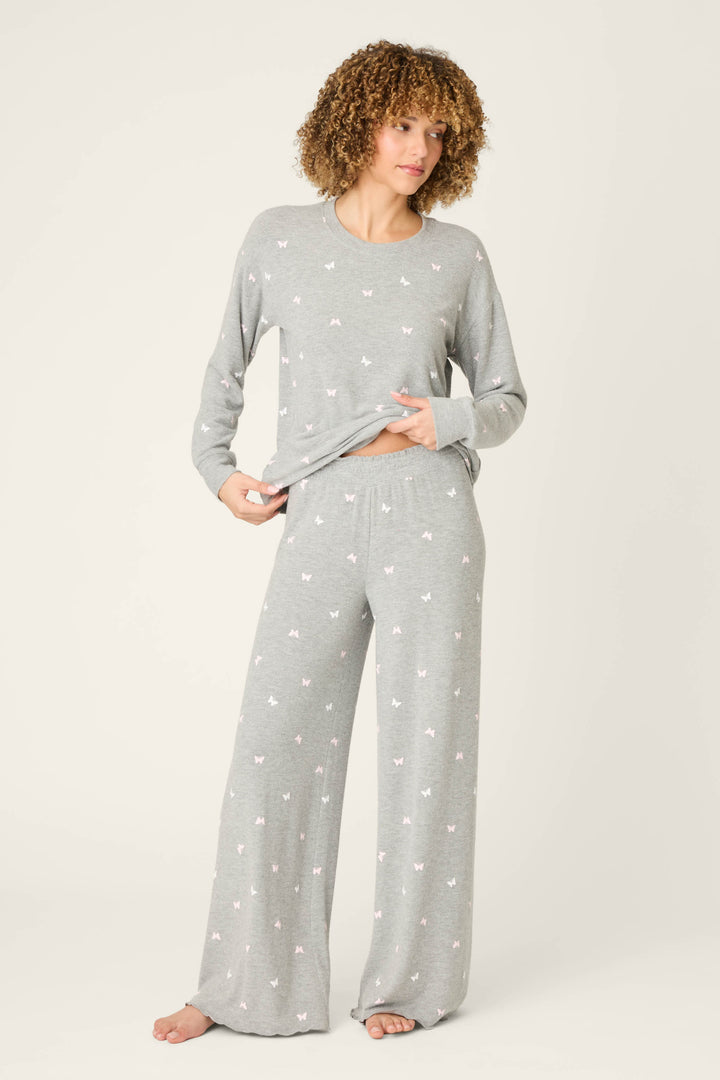 A person stands barefoot against a light background, wearing P.J. Salvage’s Peachy Mix & Match Butterfly Kisses Open Leg Lounge Pants with an elastic waistband and small white butterfly print, holding up part of their shirt to reveal their midriff.