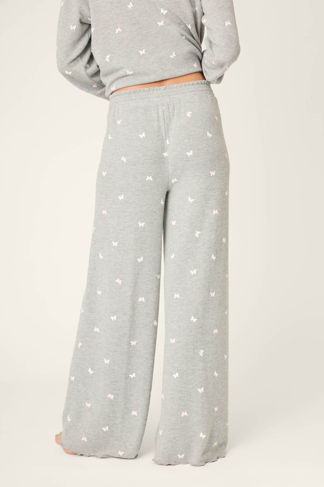 A person is seen from the back wearing P.J. Salvage Peachy Mix & Match Butterfly Kisses Open Leg Lounge Pants in light gray, featuring a white and pink butterfly print, elastic waistband, and paired with a matching top for a cozy look.