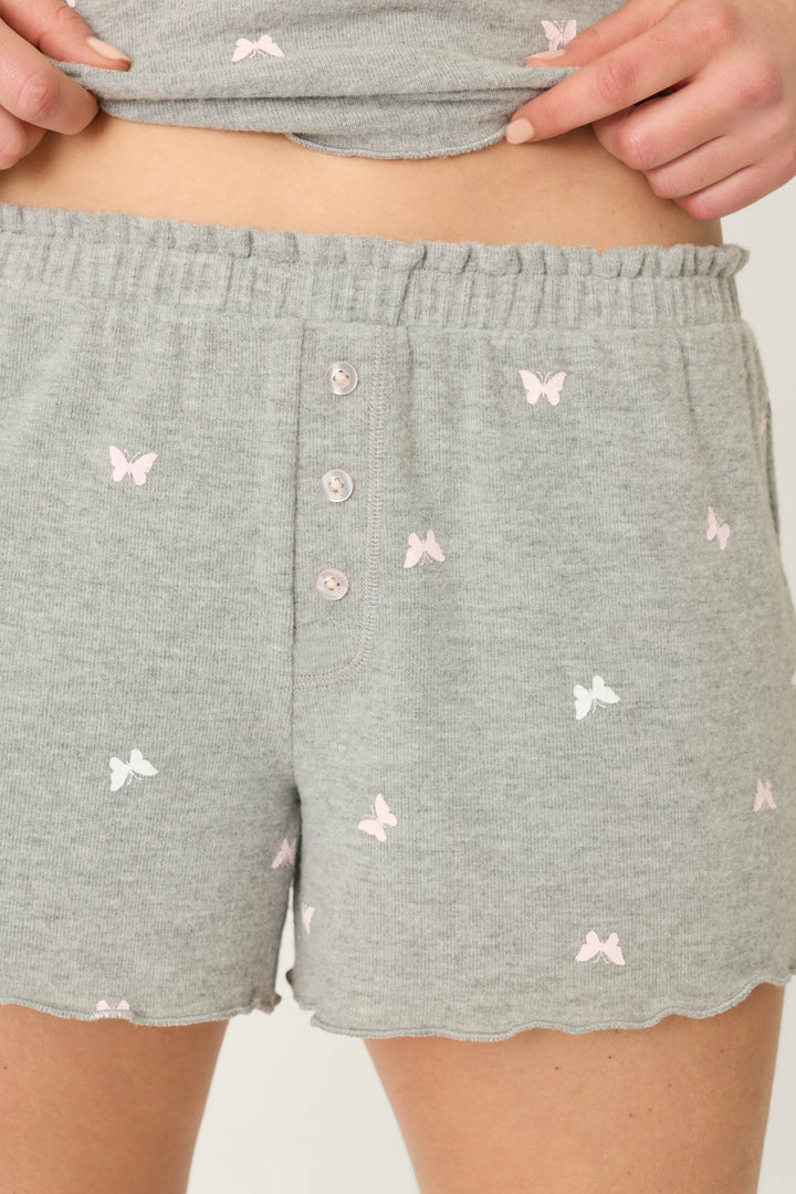 Close-up of a person wearing P.J. Salvage Peachy Mix & Match Butterfly Kisses Shorts in heather grey with pink butterflies, front buttons, and an elastic waistband, paired with a matching peachy jersey top.
