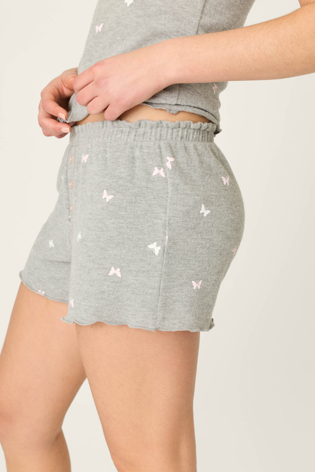 A person is wearing P.J. Salvage Peachy Mix & Match Butterfly Kisses Shorts in heather grey with a subtle pink butterfly print, paired with a matching top. The image displays a side view of the waist and upper thigh.