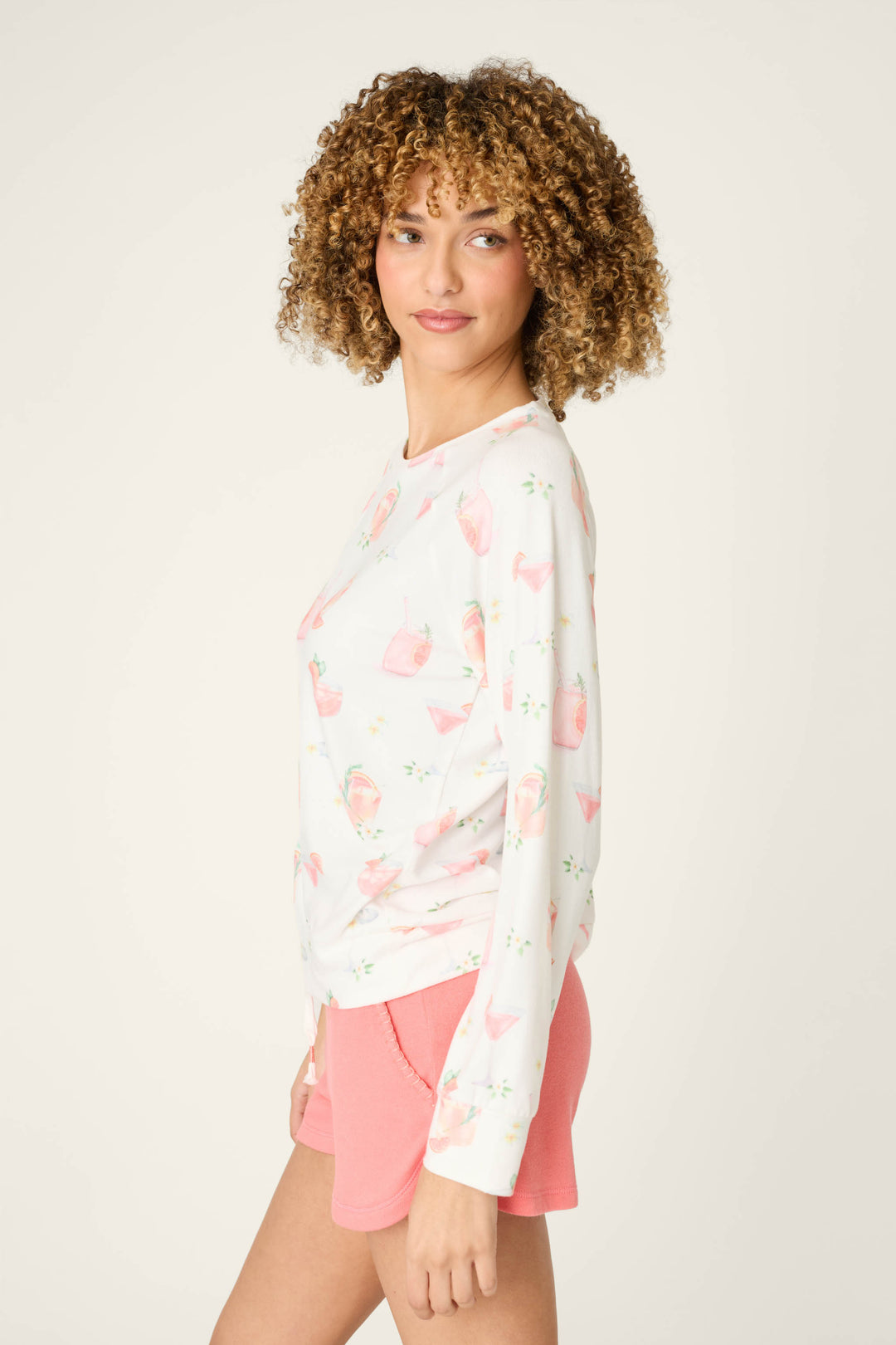 A woman with curly hair stands sideways against a plain light background, wearing the P.J. Salvage Paloma Mama Sweet Sips Long Sleeve Top in ivory with a pink and green cocktail print, paired with pink shorts.