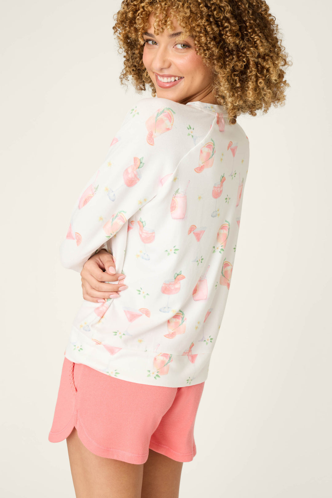 A person with curly hair smiles, wearing the P.J. Salvage Paloma Mama Sweet Sips Long Sleeve Top in ivory with a cocktail print and pink shorts, turned slightly to the side against a plain light background.