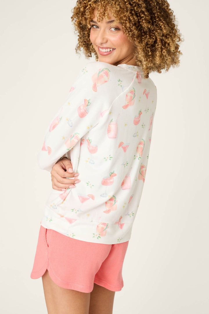 A person with curly hair smiles, wearing the P.J. Salvage Paloma Mama Sweet Sips Long Sleeve Top in ivory with a cocktail print and pink shorts, turned slightly to the side against a plain light background.