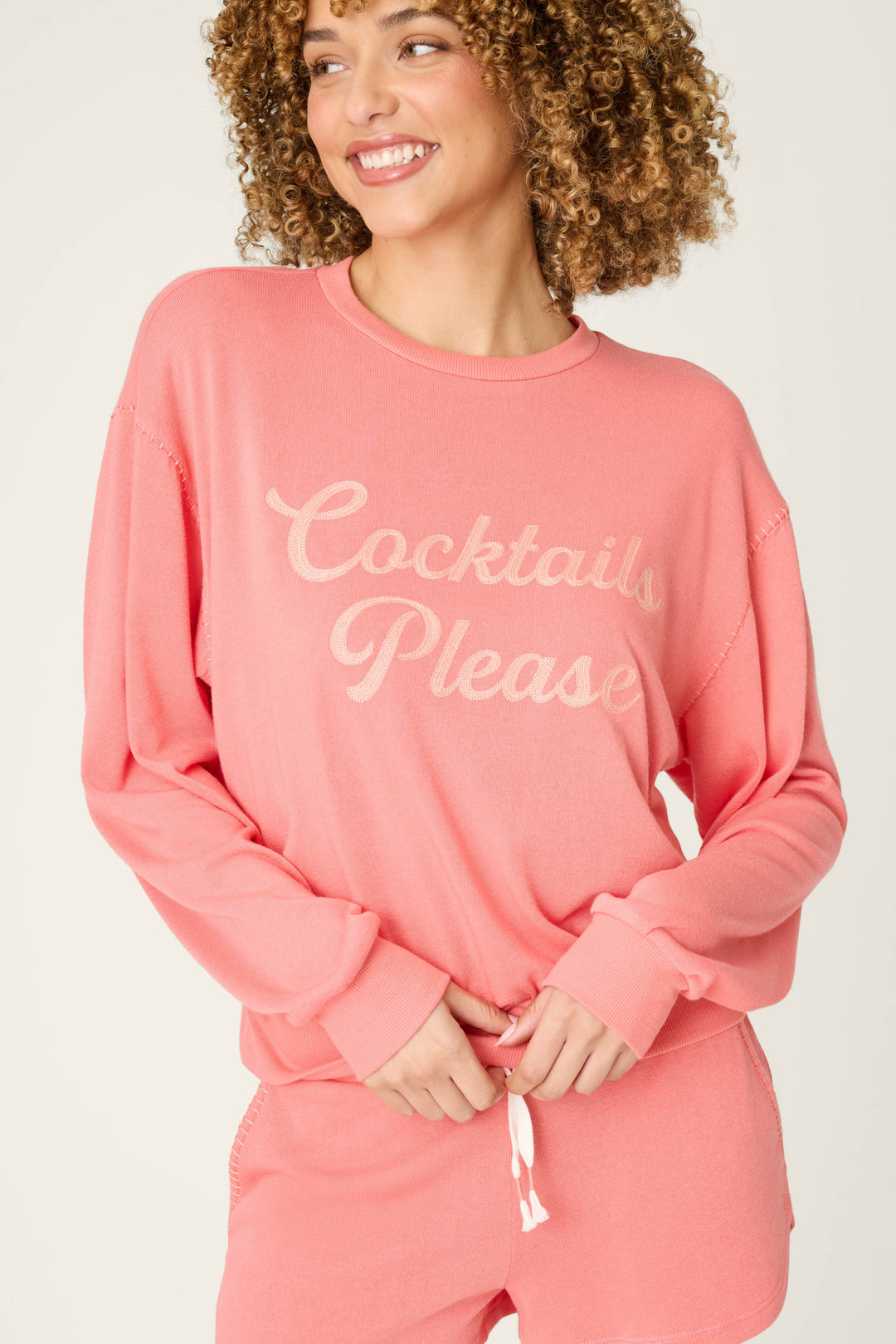 A person with curly hair wears the Paloma Mama Cocktails Please Graphic Sweatshirt by P.J. Salvage—a cozy pink French terry top with light script—while smiling and looking to the side, paired with matching shorts.